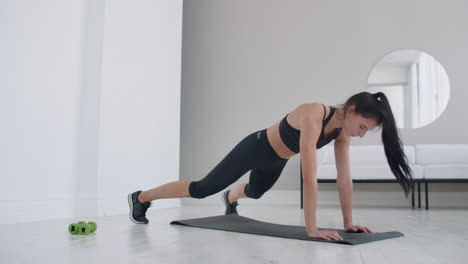 A-young-brunette-in-black-clothes-and-a-light-apartment-performs-the-plank-exercise-jumping-in-the-rest-lying-down